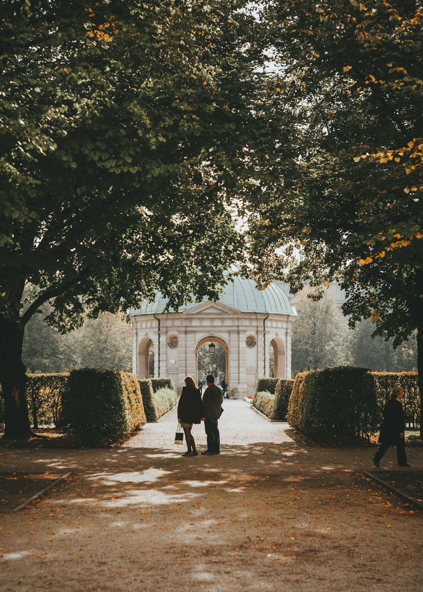 Man and woman together on an autumn pathway with a dome building behind them in Munich Hofgarten